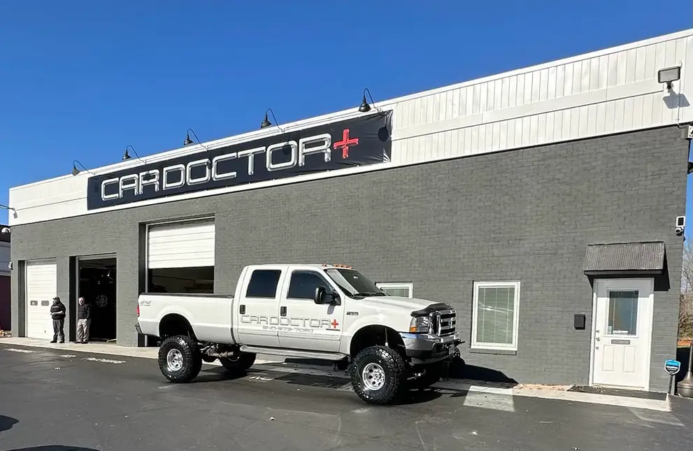 The lifted Car Doctor+ F-250 parked outside the shop's bay doors on a clear day