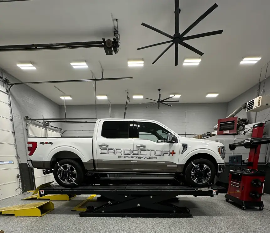 The white Car Doctor+ F-150, lettered with the shop name, raised on the scissor lift in the bay