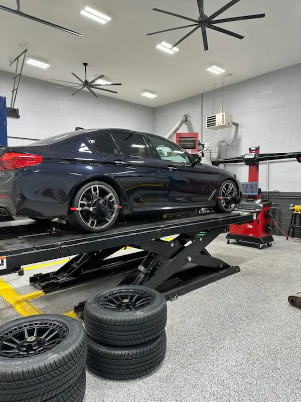 A dark BMW up on the lift with its wheels off, tires stacked on the bay floor