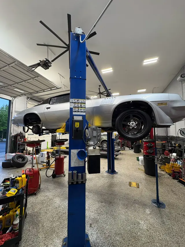 A silver Camaro raised on a blue two-post lift in the Car Doctor+ bay, drain pans and oil on the cart below
